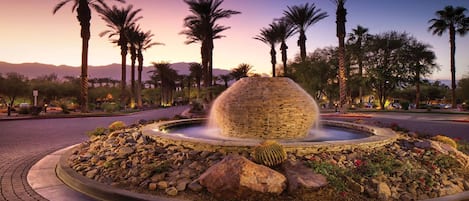 A landscaped fountain with a spherical stone structure, surrounded by palm trees and a rocky border.