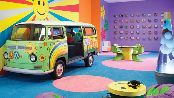 A colorful room with a green VW camper van, a yellow table, and a large smiley face mural.