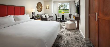 Habitación de hotel con una cama grande, ventilador de techo, televisor, escritorio con silla y vistas al exterior.