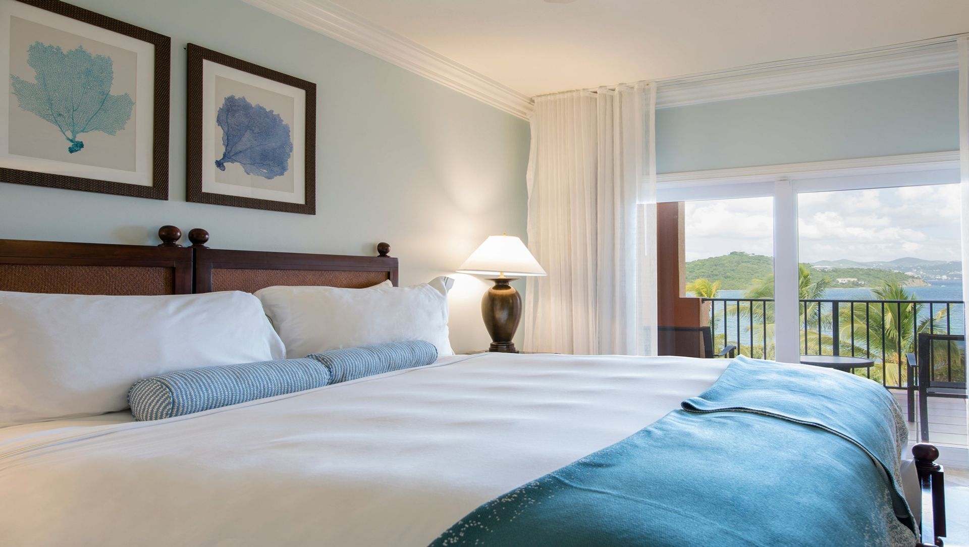 Habitación de hotel con una cama grande, dos impresiones enmarcadas de motivos botánicos en la pared, una mesita de noche con lámpara y un balcón con vistas al paisaje.
