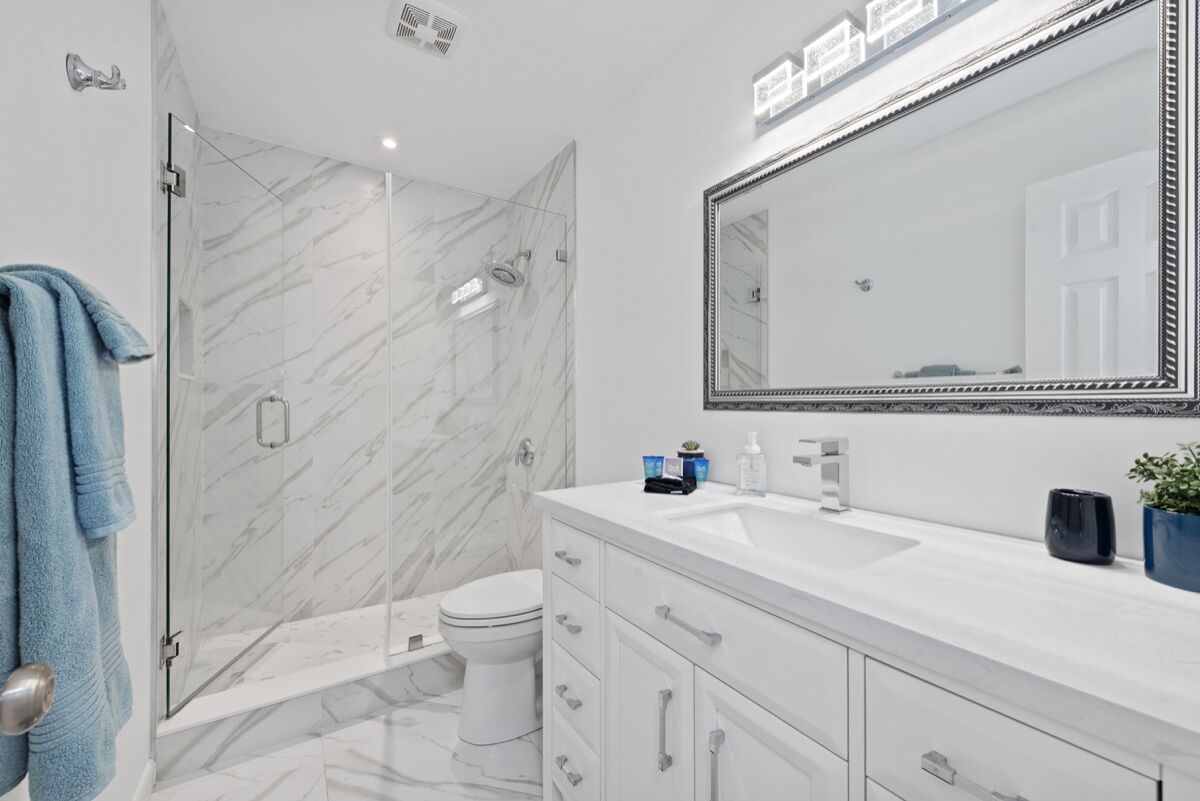 A modern bathroom with a glass shower enclosure, a white toilet, a large mirror, and a white vanity with drawers.