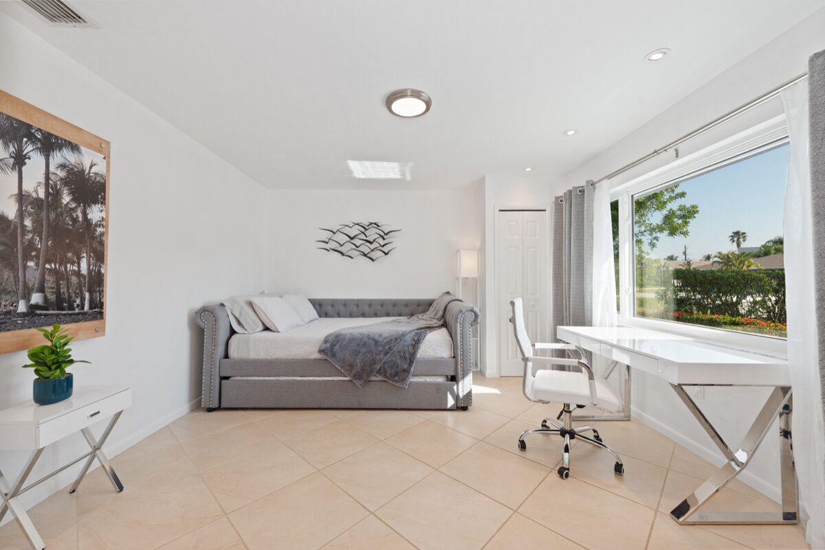 Une chambre moderne avec un canapé gris, un bureau blanc et une grande fenêtre donnant sur des palmiers et un ciel dégagé.