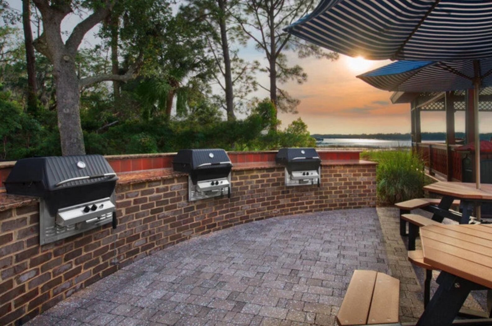 An outdoor patio with three built-in barbecue grills, a wooden table, and a covered seating area with a view of the sunset over a body of water.