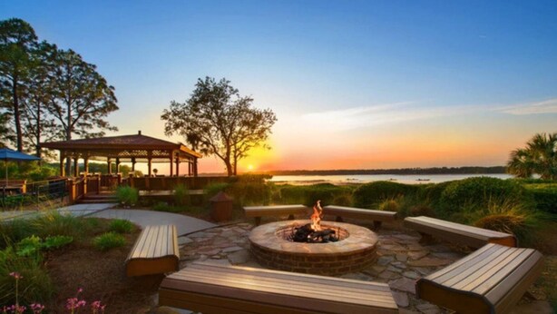 A patio area with a fire pit, wooden benches, and a gazebo overlooking a body of water during sunset.