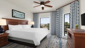 A hotel room with a large bed, two bedside tables with lamps, a ceiling fan, patterned curtains, and a view of the outdoors.