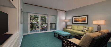 A living room with a green couch, a wooden chair, a TV, and a balcony with a view of trees.