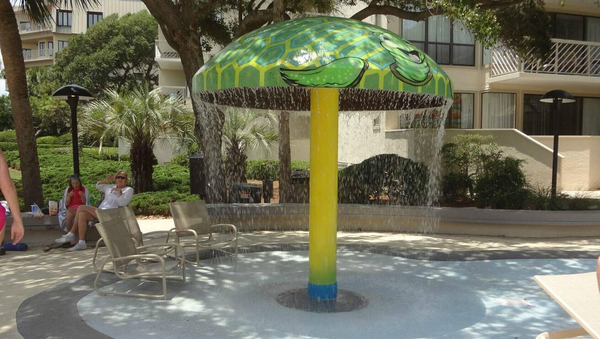 A water feature with a green and yellow umbrella-like structure, surrounded by chairs and palm trees.