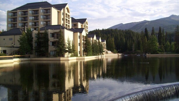 Un hotel de varios pisos con balcones, ubicado a orillas de un lago tranquilo con reflejos, rodeado de árboles y montañas.