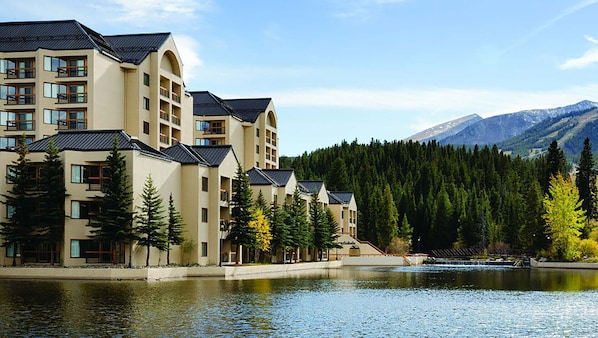 Ein mehrstöckiges Hotel mit Balkonen, einem See im Vordergrund und einem bewaldeten Berg im Hintergrund.