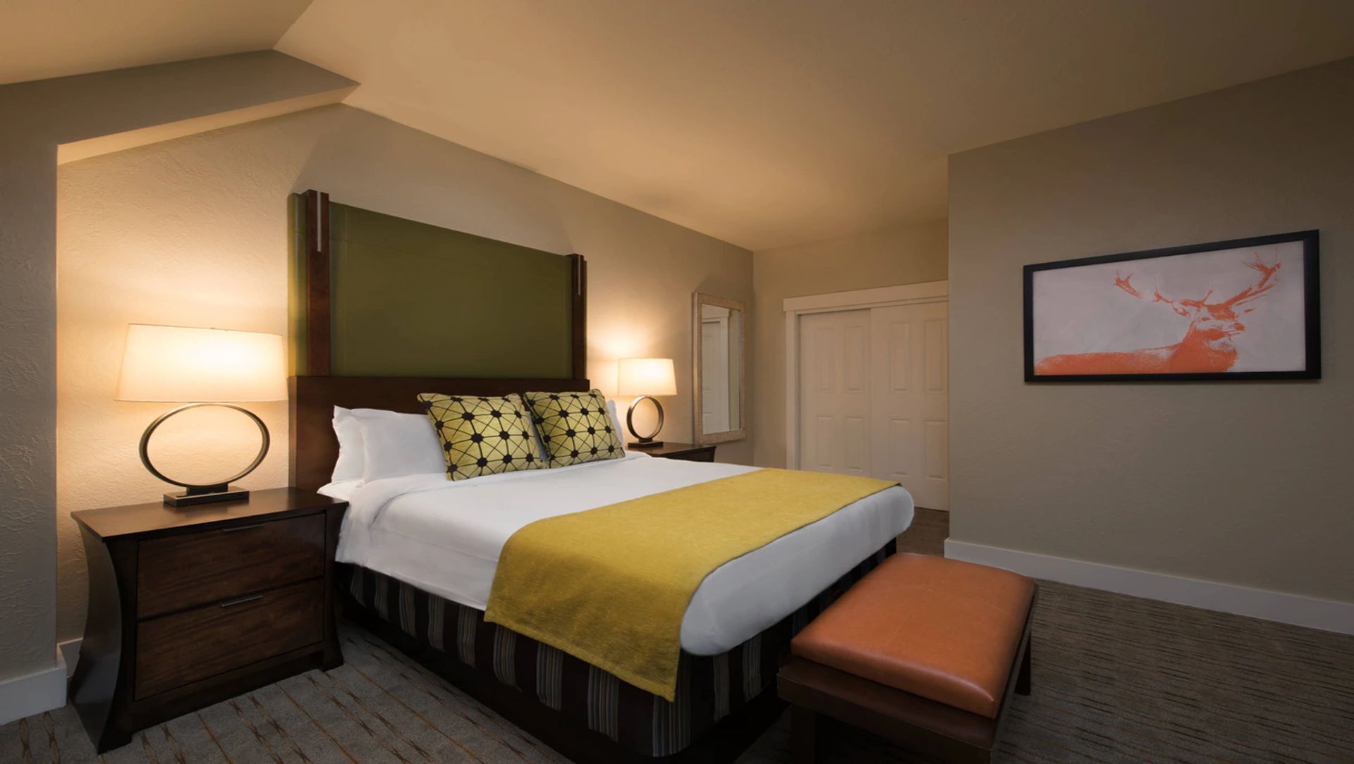 Quarto de hotel com cama, duas mesas de cabeceira com abajures, um banco, um espelho e um quadro na parede.