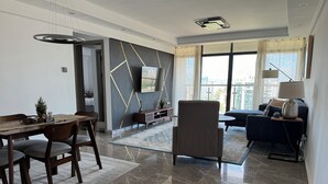 Un salon moderne comprenant un coin repas, un grand téléviseur fixé sur un mur orné d’un motif géométrique, et une vue sur la ville grâce à de grandes fenêtres.