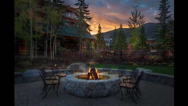 A stone fire pit with chairs around it, set in a landscaped area with trees and a building in the background.