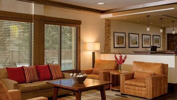 A living room with a sofa, armchairs, a coffee table, and a lamp. There are large windows with blinds, a fireplace, and framed pictures on the wall.