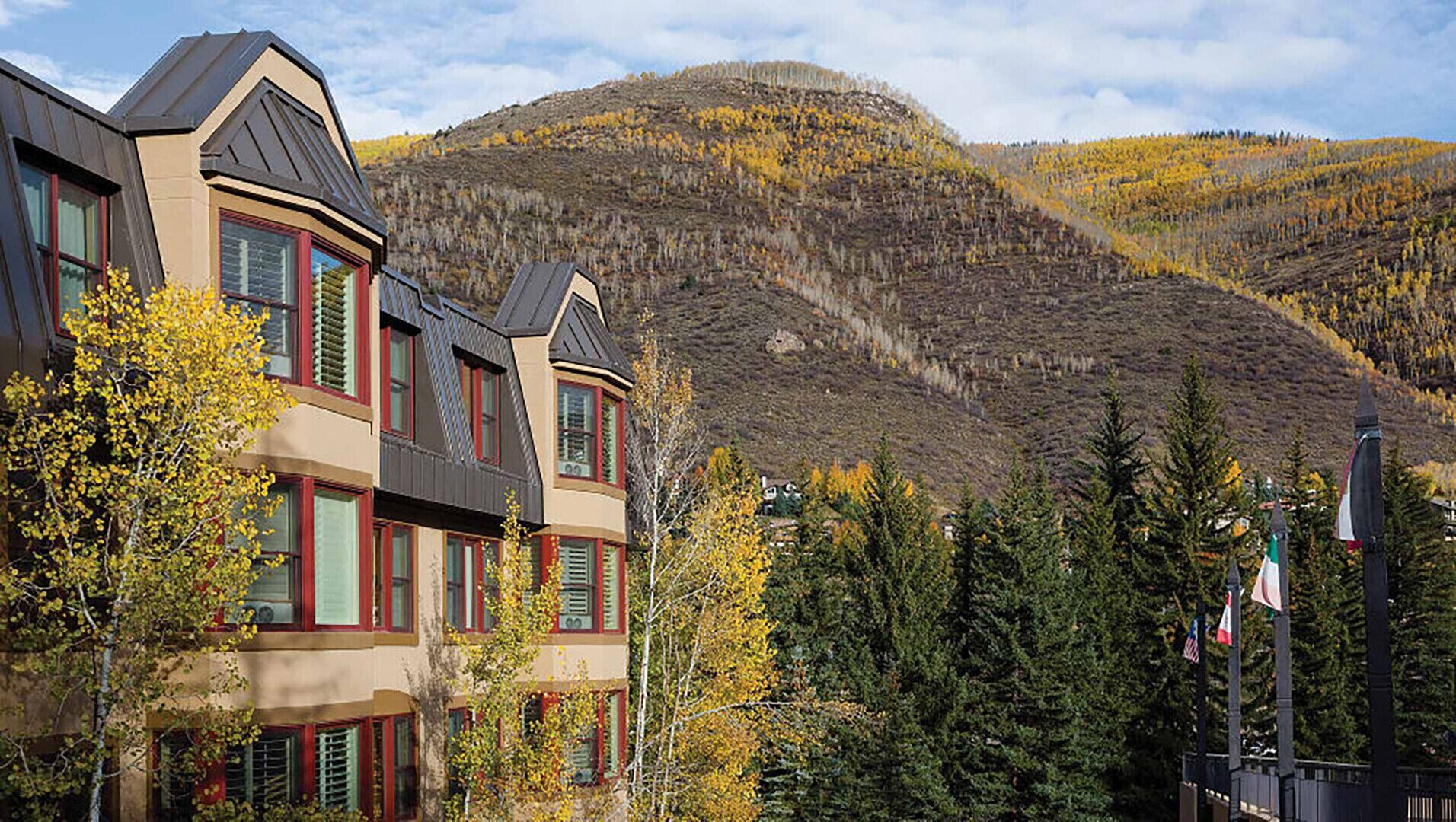 Un edificio de varios pisos con un diseño arquitectónico moderno, que cuenta con amplios ventanales y un balcón destacado. El edificio está rodeado de árboles con follaje otoñal y un telón de fondo montañoso.
