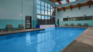 Une piscine intérieure à l’eau d’un bleu cristallin, entourée de chaises longues et dotée d’une grande baie vitrée offrant une vue sur l’extérieur.