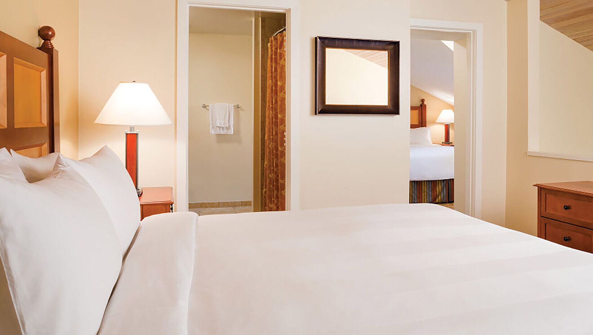 Une chambre d’hôtel avec un grand lit, une table de chevet avec une lampe, un miroir au mur et une porte donnant sur une salle de bain.