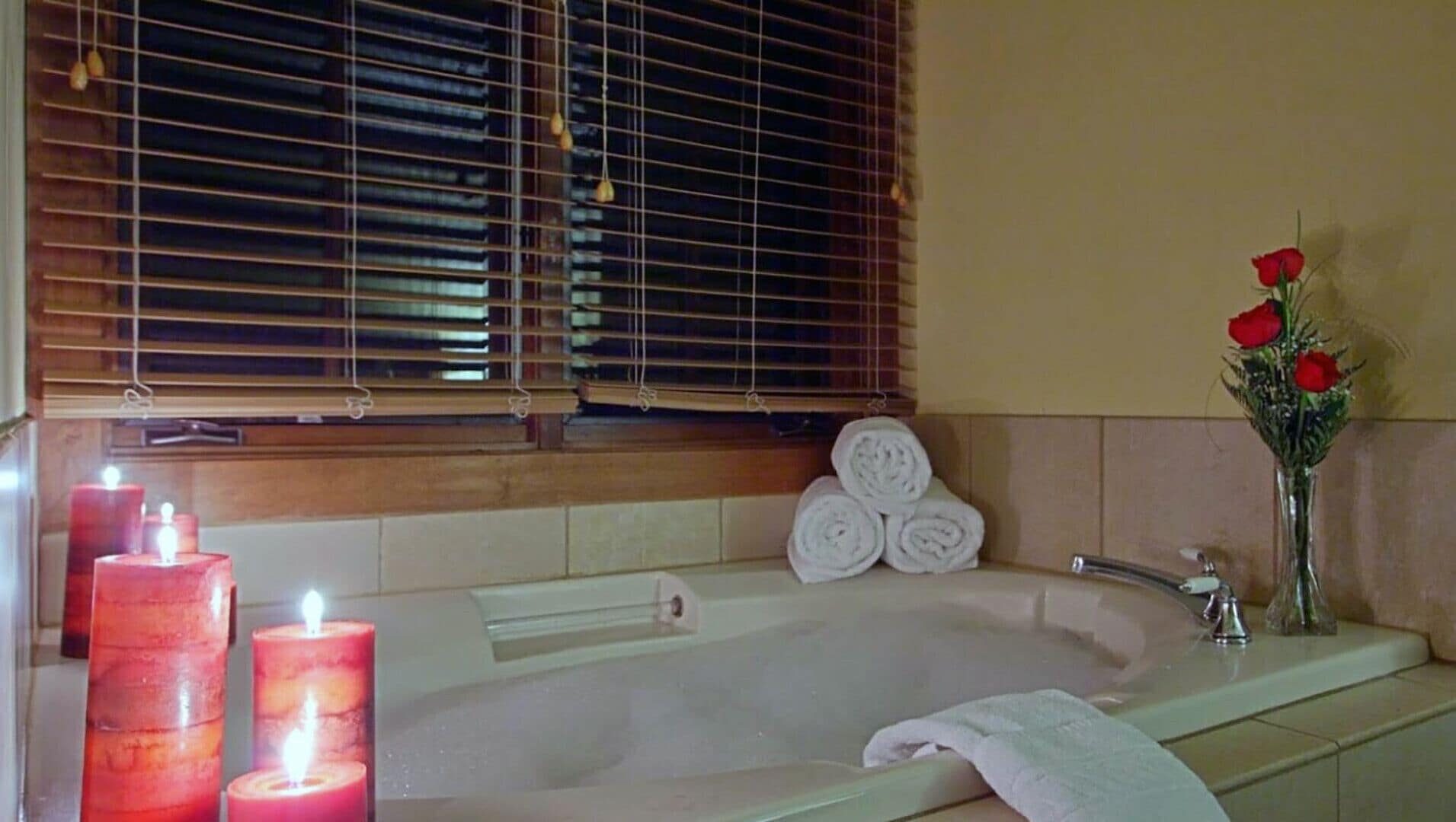 Un baño con bañera, una ventana con persianas, un florero con flores rojas y dos velas encendidas.