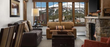 A cozy living room with a fireplace, a large window offering a mountain view, a sofa, a coffee table, and a television mounted on the wall.