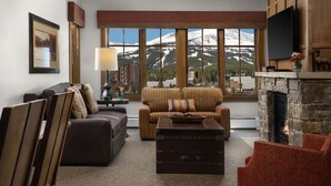 A cozy living room with a fireplace, a large window offering a mountain view, a sofa, a coffee table, and a television mounted on the wall.