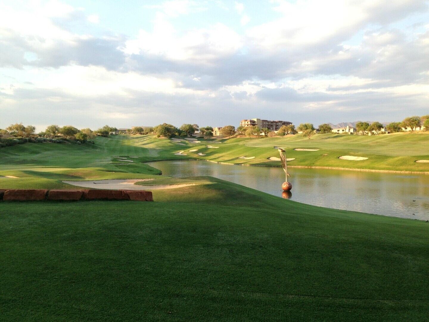 Een golfbaan met een vijver, zandbunkers en een gebouw in de verte.