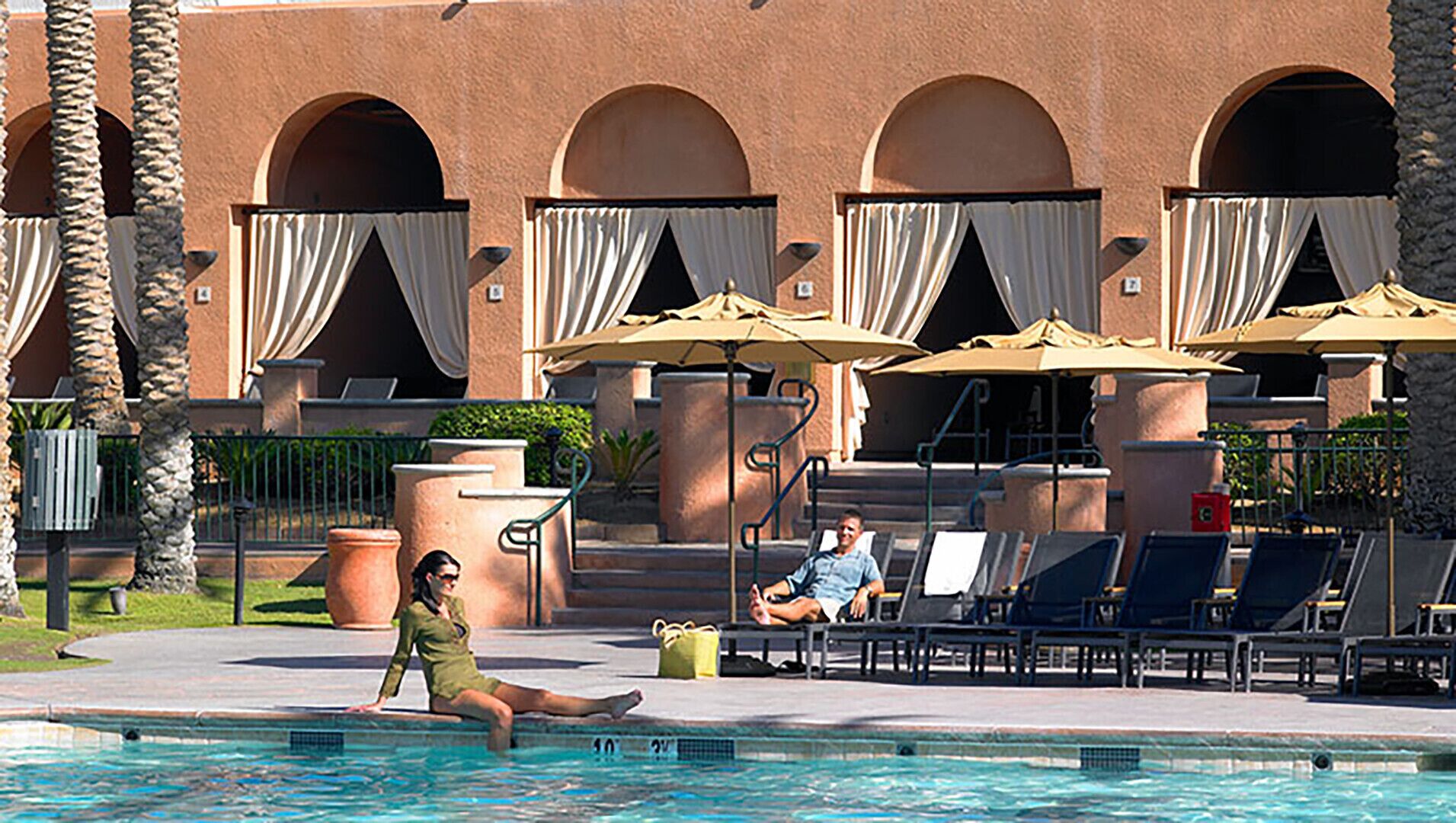 A poolside area with lounge chairs, umbrellas, and a building with arched windows.