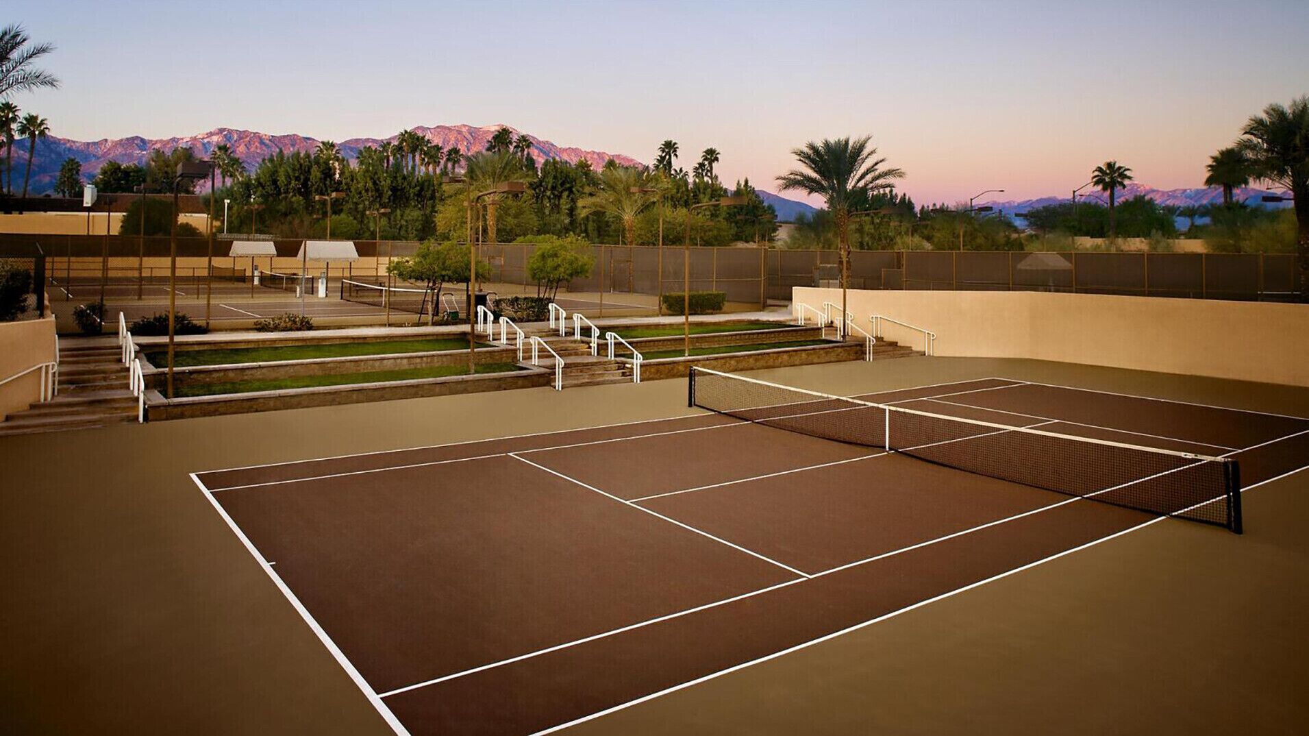 Een tennisbaan buiten met een net, omgeven door een hek en palmbomen.