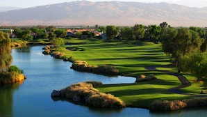 Un campo de golf con un río, casas y montañas al fondo.