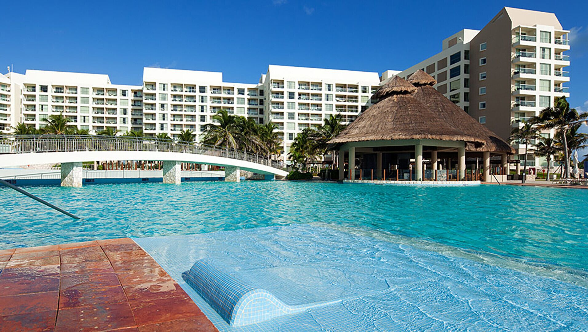 Ein Resort-Pool mit einer strohgedeckten Struktur, einer weißen Brücke und modernen Hotelgebäuden im Hintergrund.