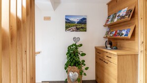 Un mueble de madera con un estante que sostiene folletos, un cuadro en la pared de un paisaje montañoso, una planta en maceta con forma de corazón, y una puerta con un panel de madera.