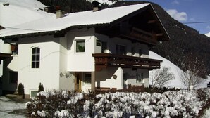 Un chalet de dos pisos con techo inclinado, balcón de madera y montañas al fondo.
