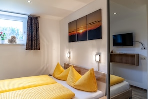 Ein Hotelzimmer mit Bett, Fernseher, Fenster mit Vorhängen und einem Sonnenuntergangsbild an der Wand.