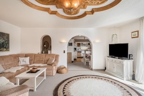A living room with a sofa, coffee table, TV, and a kitchen area visible through an archway.
