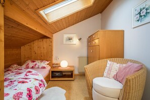 A cozy room with a sloped ceiling, a bed with floral bedding, a wicker chair, a small table with a lamp, and a radiator.