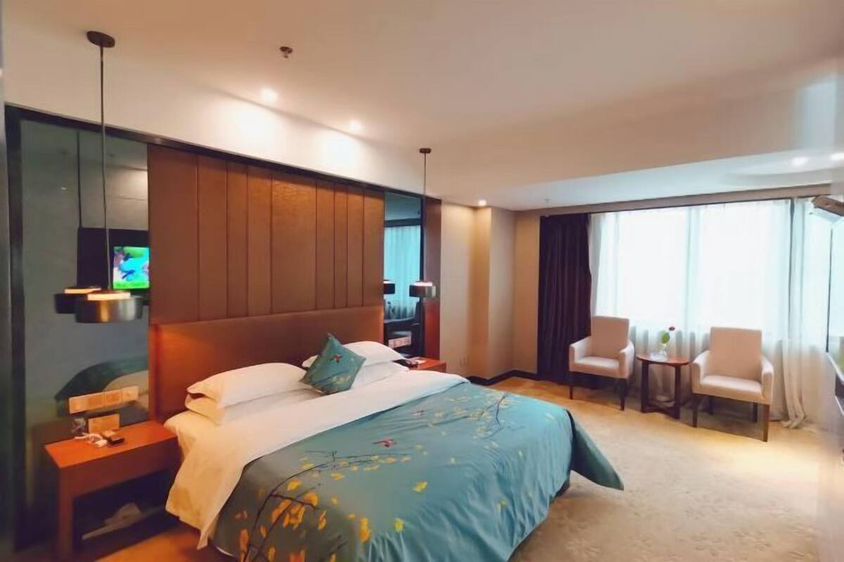 Habitación de hotel con cama, mesitas de noche, televisor, zona de estar con sillas y un gran ventanal.