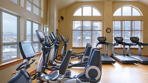 Ein Fitnessraum mit mehreren Cardiogeräten, großen Fenstern und einer Uhr an der Wand.