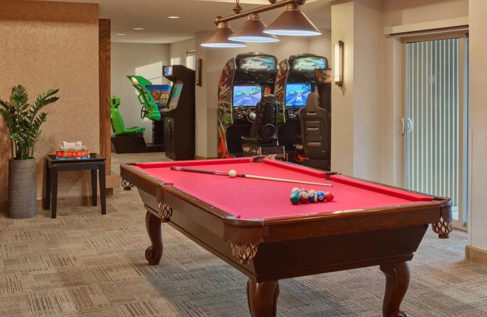 Une table de billard recouverte d’un drap de feutre rouge, entourée d’un sol recouvert de moquette. Au fond, on aperçoit deux bornes d’arcade et une plante en pot.