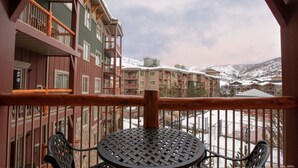 A balcony with a metal table and chairs, overlooking a snow-covered mountain area with multi-story buildings.