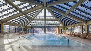 An indoor swimming pool with a glass ceiling, lounge chairs, and a view of the outside.