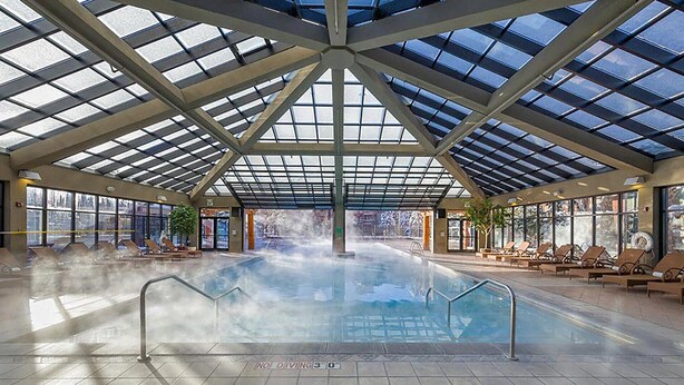 An indoor swimming pool with a glass ceiling, lounge chairs, and a view of the outside.