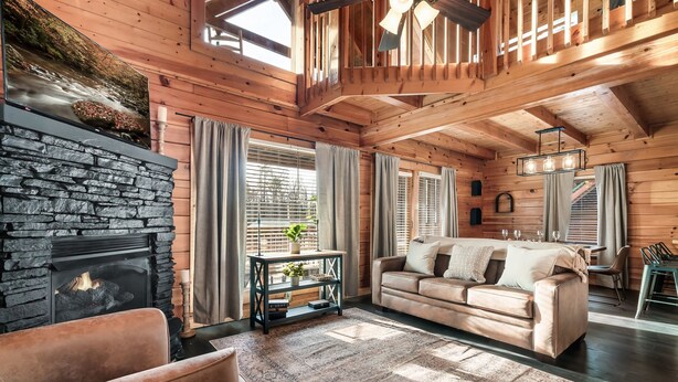 Ein gemütliches Wohnzimmer mit Kamin aus Stein, einem beigen Sofa und einem großen Fenster mit Vorhängen.