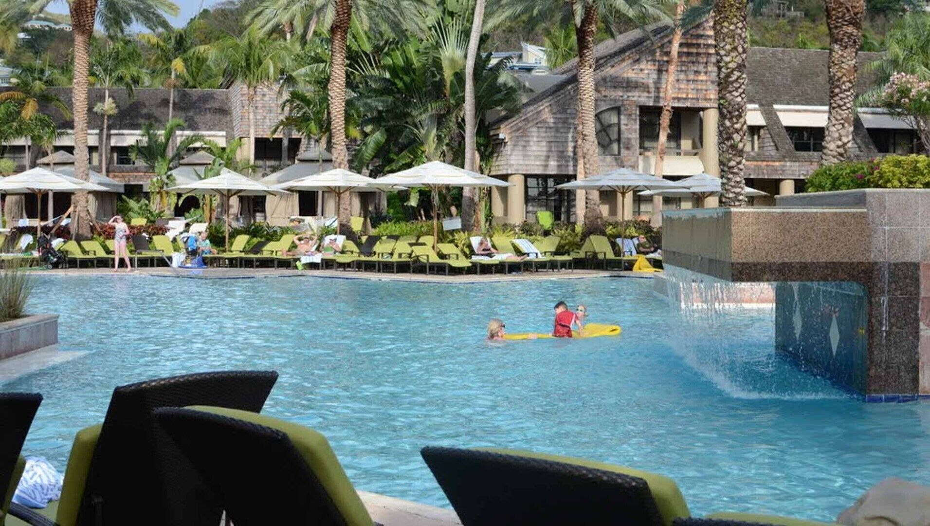 A pool area with lounge chairs and umbrellas, surrounded by palm trees and a building with a stone facade.