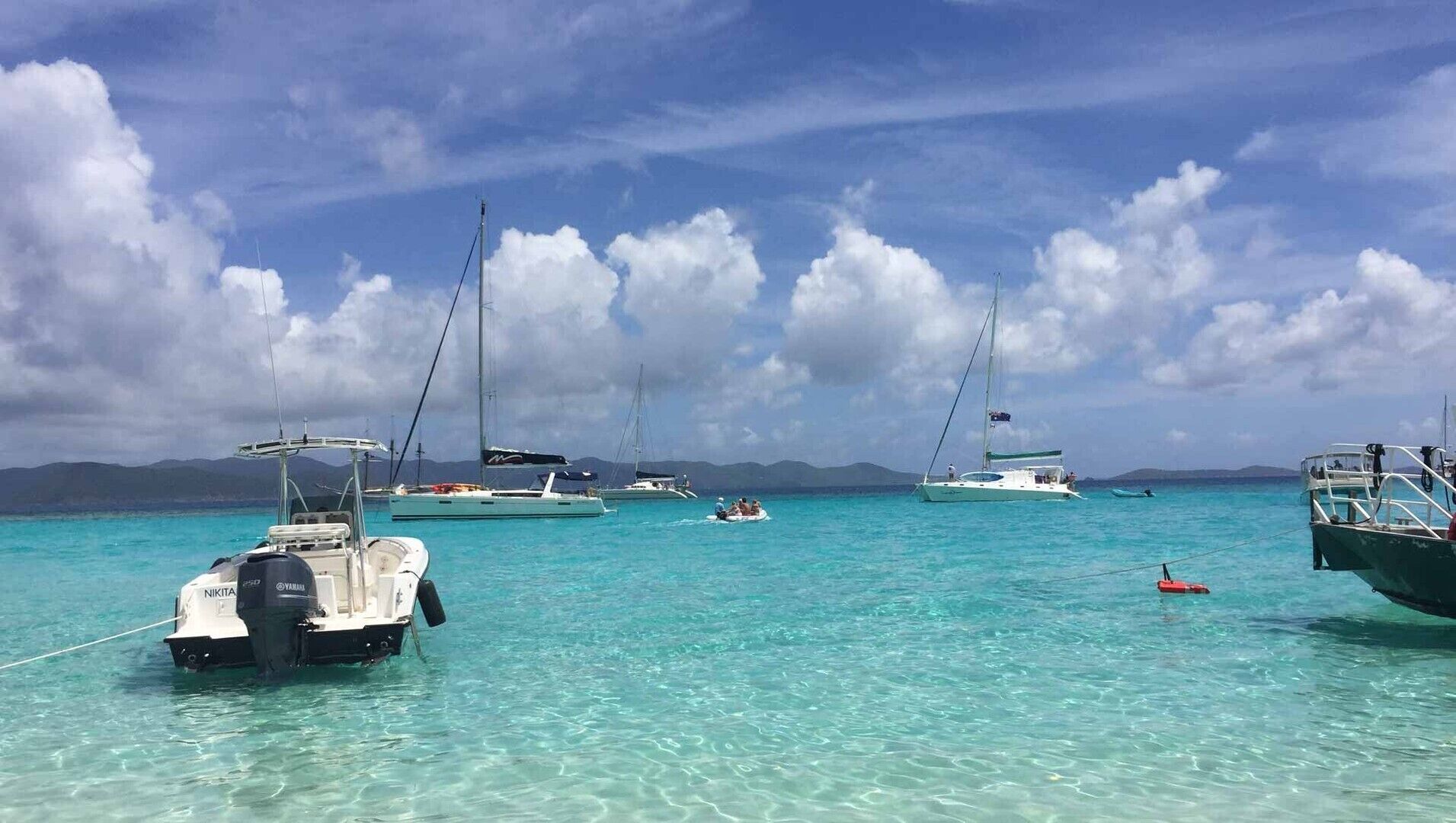 Ein Yachthafen mit mehreren Booten, die in klarem, türkisblauem Wasser vor Anker liegen, mit Bergen im Hintergrund.