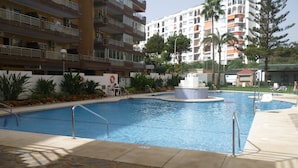 A swimming pool with a fountain, surrounded by stone tiles and greenery, with multi-story buildings in the background. - Modern 1 Bed Seafront Apt Los Boliches (Fuengirola)