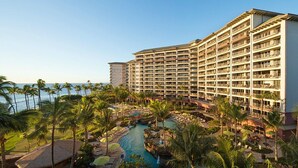 A large hotel with multiple floors, a pool area with lounge chairs, and a view of the ocean.