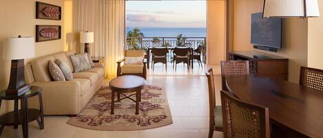 Un cuarto de hotel con sofá, una mesa de centro, una mesa de comedor con sillas, televisor y balcón con vista al océano.