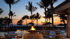 A poolside lounge with wicker chairs, a fire pit, and a view of the ocean at sunset.