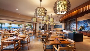 A spacious restaurant with wooden tables and chairs, large hanging lamps, and a view of the ocean.