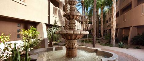 Un patio con una fuente central, rodeado de palmeras y cactus, y edificios de varios pisos con balcones.