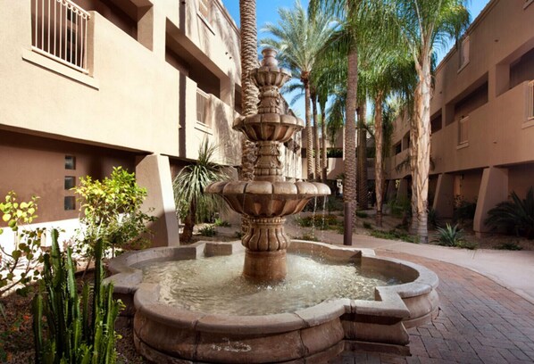 A courtyard with a central fountain, surrounded by palm trees and cacti, and adjacent multi-story buildings with balconies.