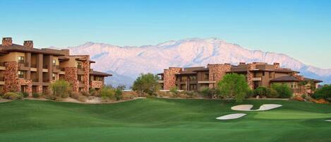 A golf course with two buildings featuring stone facades and multiple balconies, set against a mountainous backdrop.
